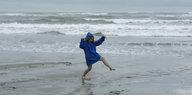 Ein Mädchen in blauer Regenjacke hüpft mit nackten Beinen durch die Brandung, die den trüb-grauen Himmel spiegelt