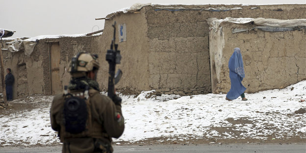 Ein afghanischer Soldat steht mit dem Rücken zum Betrachter und mit Waffen im Arm vor einem verschneiten Grundstück, über das eine Frau in blauem Gewand geht