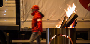 Ein Mann in roter Jacke und rotem Käppi geht hinter einer brennenden Feuertonne entlang