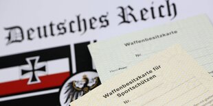 Ein Blatt Papier mit einer Abbildung der Fahne des Deutschen Reiches und des Eisernen Kreuzes. Auf dem Blatt liegen zwei Waffenscheine.