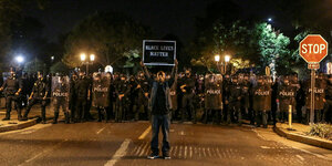 Ein schwarzer Mann steht vor einer Reihe von Polizisten mit einem Schild "Black Lives Matter"