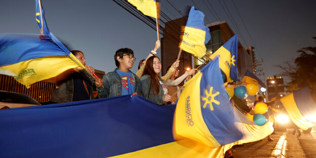 Menschen stehen jubelnd entlang einer Strasse, sie halten ein blau-gelbes Transparent und blau-gelbe Fahnen hoch