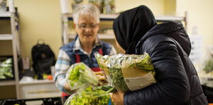 Eine Frau steht hinter Gemüsekörben, eine andere hält einen Beutel Salat im Arm