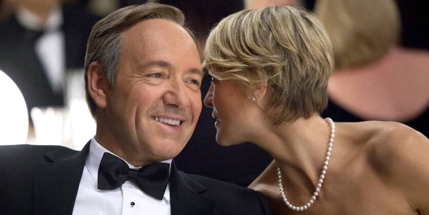 Francis Underwood (Kevin Spacy) und Claire Underwood (Robin Wright) sitzen gemeinsam an einem Tisch. Sie flüstert ihm etwas ins Ohr,