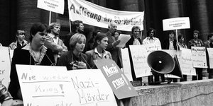 Mehrere Menschen sitzen und stehen an einer Mauer. Sie tragen Schilder um den Hals und halten Banner in die Höhe, auf denen sie die Veruteilung von Nazis und die Auflösung von SS-Organisationen fordern