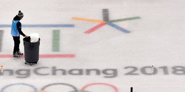 Eine Person schiebt eine Tonne über die Eisfläche an dem Logo der Olympischen Spiele vorbei
