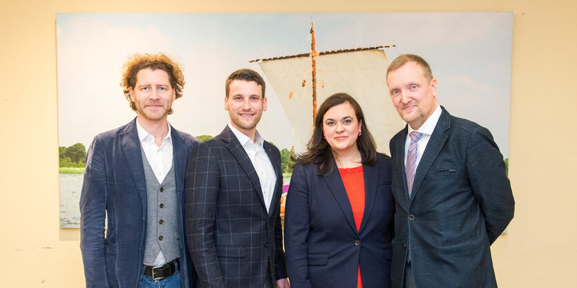 Drei Männer und eine Frau stehen vor einem Bild mit einem Kreuz. Alle vier Personen tragen Sakkos.