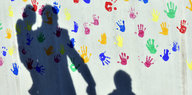 Schatten einer Person, die ein Kind auf dem Arm hat und ein Kind an der Hand hält. Im Hintergrund bunte Handabdrücke von Kindern.