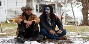 Zwei Männer sitzen auf dem Boden vor einem Haus. Einer hat einen Hut mit gebogener Krempe, der andere ein blau-weiß bemaltes Gesicht