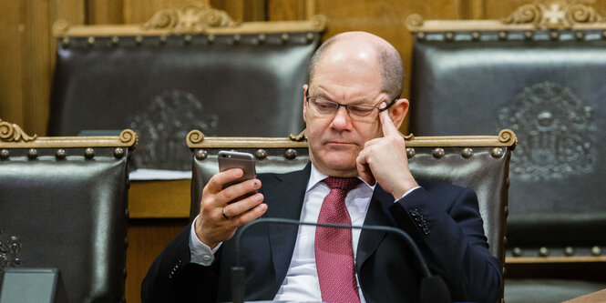 Ein Mann mit Glatze, Brille und Anzug sitzt auf einem Stuhl mit Lederpolstern und blickt missmutig auf sein Handy.