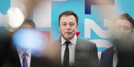 Elon Musk bei einer Messe. Zwei Männer neben ihm unscharf