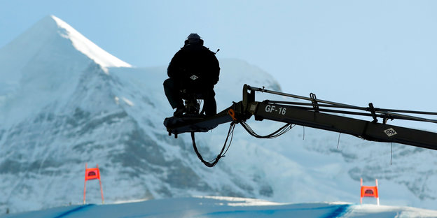 Ein Kameramann der sitzt auf einem Träger hoch über verschneiten Hügeln, die mit Skirenntoren bestückt sind