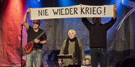 Zwei mittelalte Männer halten ein Plakat, auf dem steht: "Nie wieder Krieg!" Zwischen den Männern steht eine 93-Jährige Frau hinter einem Notenständer.