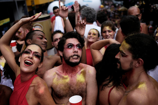 Menschen feiern auf einer Straßenparade. Im Vordergrund zwei Männer mit nacktem Oberkörper und goldener Glitzerfarbe auf der Brust und eine Frau in einem rotem Oberteil.