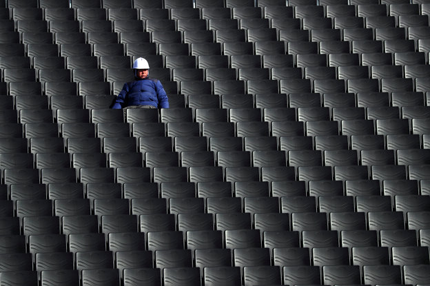 Dutzende leere Stühle auf einer Tribühne. Nur ein Mann sitzt auf einem der hinteren Ränge in blauem Anzug und weißem Helm.
