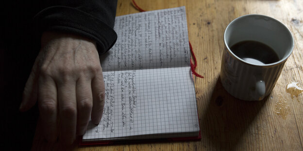 Ein Notizbuch ist aufgeklappt, die Hand einer älteren Frau liegt daneben, eine Kaffeetasse steht davor
