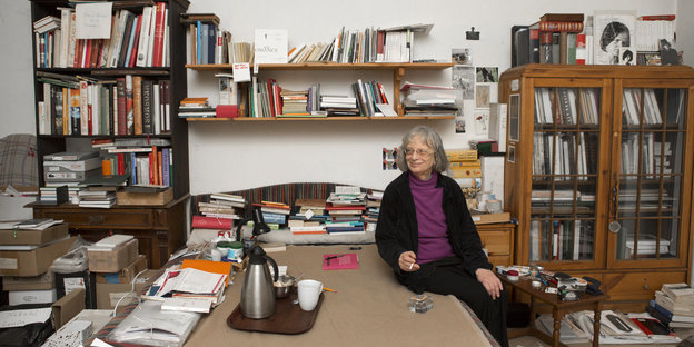 Eine ältere Frau sitzt auf einem Bett, umgeben von Bücherregalen und Bücherstapeln Eine ältere Frau sitzt auf einem Bett, umgeben von Bücherregalen und Bücherstapeln