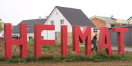 Riesige rote Buchstaben stehen auf einer Wiese vor einer Eigenheimsiedlung