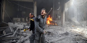 Zwei Männer stehen vor einem zerstörten Gebäude, im Hintergrund ist Rauch und Feuer, einer der Männer ruft etwas