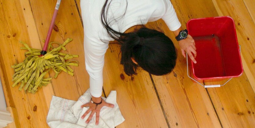 Eine Frau mit schwarzen Haaren kniet auf dem Boden. Neben ihr sind ein Wischmob, ein Lappen und ein Putzeimer.