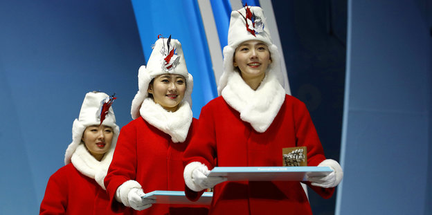 Drei Frauen tragen jeweils ein Tablett. Sie haben eine rote Jacke und weißen Schal und Mütze an.