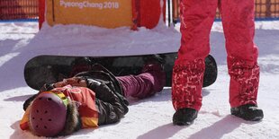 Silvia Mittermüller liegt an ihr Snowboard geschnallt auf dem Schnee. Neben ihr steht jemand in roten Skihosen