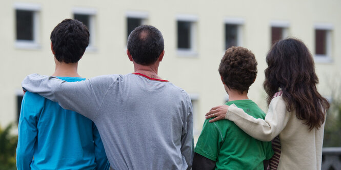 Vier Menschen sitzen nebeneinander. Zwei ältere haben den jüngeren einen Arm um die Schultern gelegt. Man sieht sie von hinten