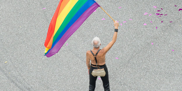 Ein grauhaariger Mann mit freiem Oberkörper und ausgeschnittener Lederhose schwenkt eine Regenbogenfarbe.