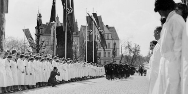 Weiß gekleidete Frauen stehen unter Hakenkreuz-Fahnen am Straßenrand. Auf der Straße marschieren dunkel gekleidete Männer.
