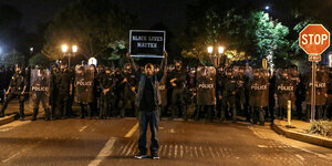 Ein farbiger Mann mit einem Protestplakat steht vor einer Wand aus Polizisten. Auf dem Schild steht Black lives matter