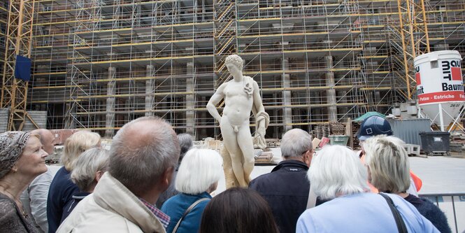 Vor einer Baustelle steht eine Statue. Menschen blicken darauf