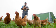 Hühner im Gras, im Hintergrund steht ein Bauer, der eines auf dem Arm hält
