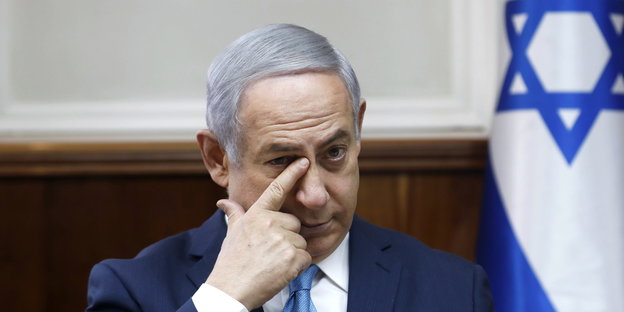 ein Mann fasst sich mit dem rechten Zeigefinger in den rechten Augenwinkel, neben ihm eine israelische Fahne