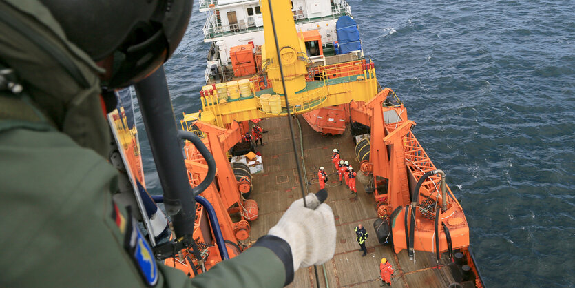 Auf einem Schiffsdeck stehen Männer in orangen Overalls. Auf einem Mast über ihnen steht ein Polizist mit olivgrüner Jacke.