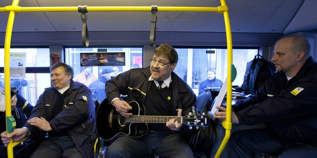 Eine BVG-Angestellter spielt in einem Bus Guitarre, zwei Kollegen sitzen danebe