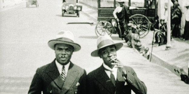 Zwei Männer mit Hut im Bildvordergrund - auf einer Straße
