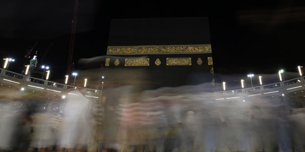 Im Vordergrund verschwommene Silhouetten vorbeilaufender Menschen, im Hintergrund die Kaaba vor dem schwarzen Nachthimmel.