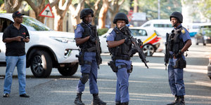 Drei bewaffnete Polizisten stehen auf einer Straße, im Hintergrund sieht man ein Auto und einen Zivilisten