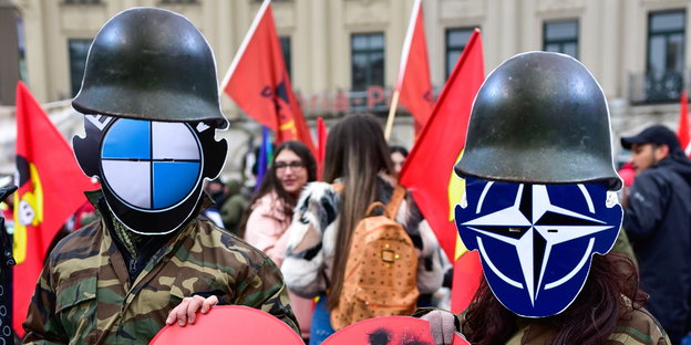 Zwei als Soldaten verkleidete Menschen, vor den Gesichtern ein BMW- bzw. ein Nato-Logo