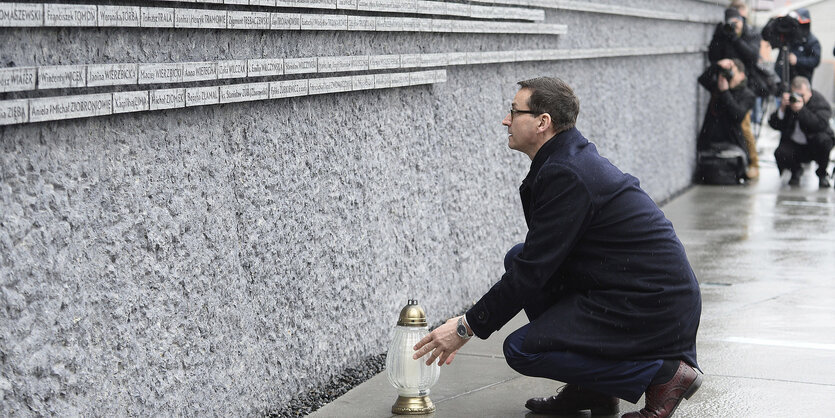 ein Mann kniet vor einer grauen Gedenkmauer nieder und berührt eine Lampe