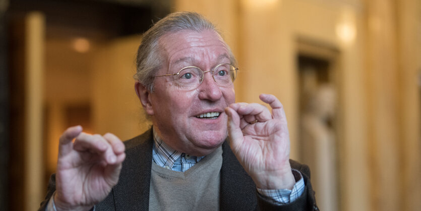 Ein Mann mit grauen Haaren und goldener Brille hebt die Hände und drückt Daumen und Zeigefinger zusammen.