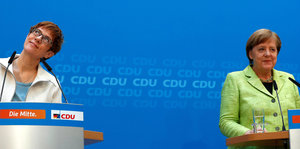 am linken Bildrand Annegret Kramp-KArrenbauer, am rechten Angela Merkel - sie stehen jeweils hinter einem Pult
