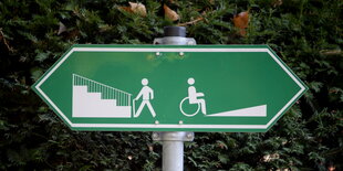 ein grünes Schild, das Pfeile nach links und rechts hat, auf der linken Seite eine Treppe und das Piktogramm eines gehenden Menschen, auf der rechten Seite eine Rampe und ein Rollstuhlfahrer