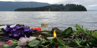 Blumen und Kerzen liegen im Regen am Ufer eines Sees, im Hintergrund ist eine Insel zu sehen