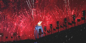 ein rotes Feuerwerk über einem Stadion, in der Mitte brennt ein monumentales olympisches Feuer