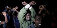 Mehrere afrikanische Männer halten bei Nacht freudestrahlend ihre Arme über dem Kopf gekreuzt.