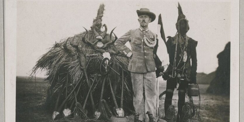 Ein weißter Mann mit dreckiger Uniform steht neben einem schwarzen Mann mit traditioneller Kleidung. Im Hintergrund eine Hütte mit Geweihen.