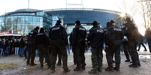 Polizisten und Polizistinnen stehen mit dem Rücken zur Kamera auf einem Platz. Im Hintergrund sieht man ein Stadion