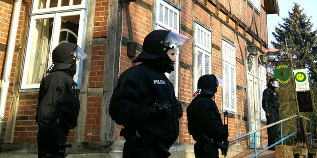 Vier vermummte Polizisten stehen in schwarzer Kampfmontor vor einem alten Backsteinhaus.