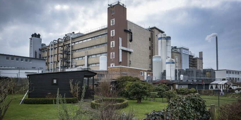 Ein Gartenhaus aus Holz steht auf einem kurz gemähten Rasen. Dahinter ein grau-braunes Fabrikgebäude mit Silos, Rohren und einem rauchenden Schlot.